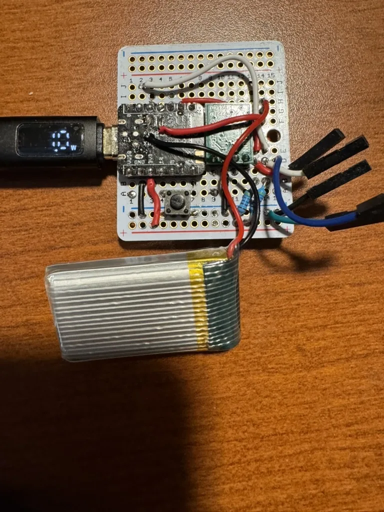 A XIAO module soldered to a small perfboard with a boost converter and a sync button, a pouch cell beside it and a USB power meter on the plug
